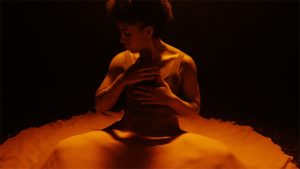A dancer is bathed in deep orange light in a black box space. They sit cross-legged on the floor, their hands on their chest. The dress they wear is splayed around them in a circle on the floor.