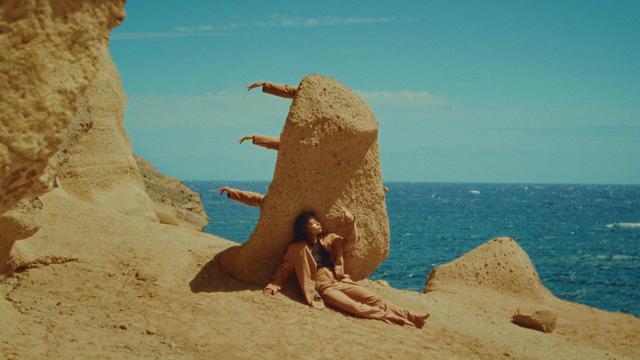 A dancer in a tan colored suit sits on a rocky cliff in front of a large, tan rock, her arm extending in front of her, palm out towards the sun. Three additional arms stick out from behind the rock. The ocean is a deep blue in the distance.