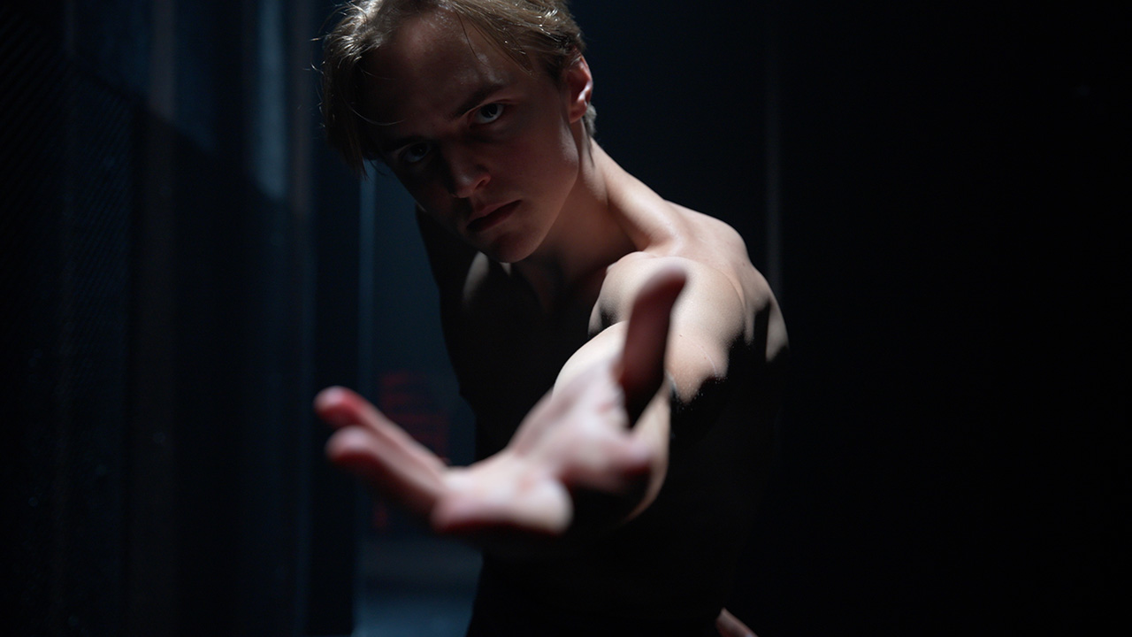 A shirtless dancer reaches an arm towards the camera, his brows furrowed. One light shines on him from above, the rest of the image shrowded in darkness.