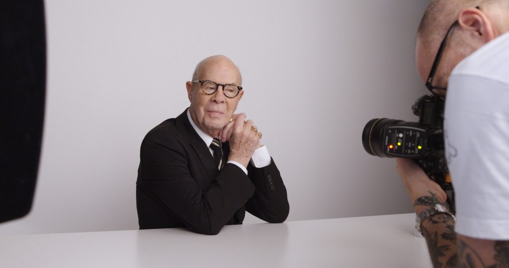 Hans van Manen, a 90-year-old white man, wears a black jacket, white collard shirt, and striped tie. He sits posing for the camera in a portrait-style photoshoot. He balances his head on his hands and wears gold rings with tortoise shell glasses.