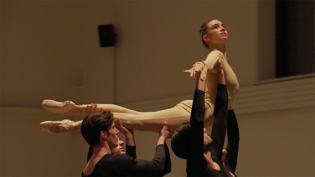 Four dancers in black costumes lift a ballerina in a beige leotard. The ballerina is extended horizontally, gazing upward, while the dancers support her gracefully in a well-lit dance studio.