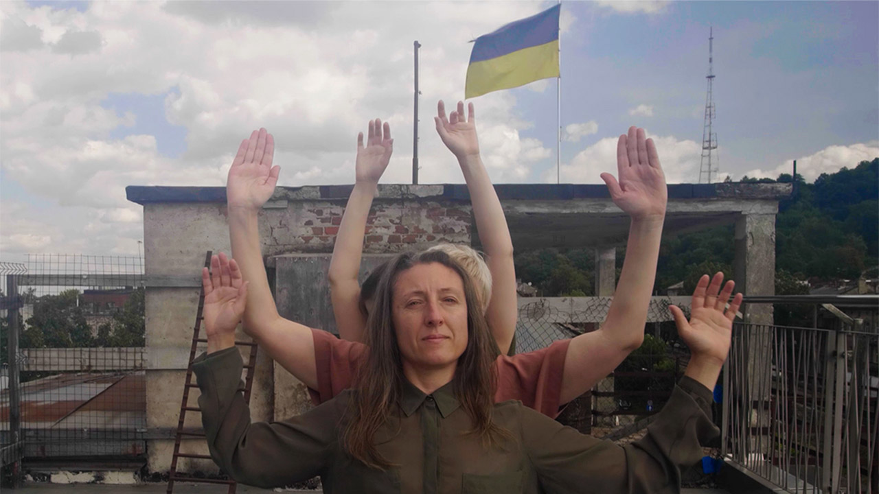 A group of dancers stand tightly aligned in line on a rooftop, their arms rasied and layered behind one another to create the illusion of a single figure with multiple arms. In front, a dancer with long brown hair wearing an olive-green military style shirt holds their hands up in a gesture of surrender, while a Ukranian flag waves in the background.